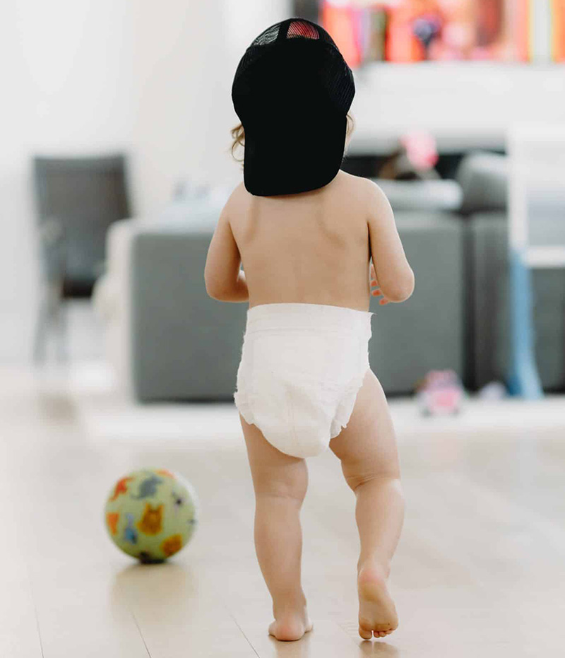 A toddler walking indoors with a black cap and diaper, home living room background.